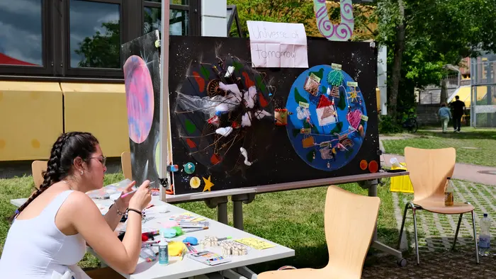 Eine Person sitzt an einem Tisch und arbeitet an Bastelmaterialien. Im Hintergrund ist eine Station mit zwei großen, bunten Plakaten, die fantasievolle Welten darstellen. Über den Plakaten hängt ein Schild mit der Aufschrift „Unseres Tomorrows“. Der Ort ist im Freien, umgeben von Gras und Bäumen.