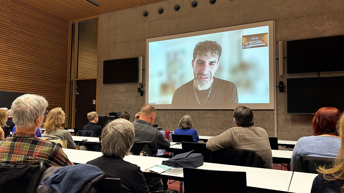 In einem Vorlesungssaal lauschen die Teilnehmenden der Poetikdozentur den Worten von Necati Öziri, der mittels einer Videoübertragung live zugeschaltet ist