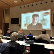 In einem Vorlesungssaal lauschen die Teilnehmenden der Poetikdozentur den Worten von Necati Öziri, der mittels einer Videoübertragung live zugeschaltet ist