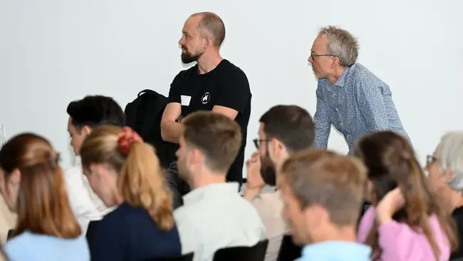 Zwei Männer stehen in einer Gruppe von Menschen. Der Mann links trägt ein schwarzes T-Shirt und schaut aufmerksam nach vorne, während der Mann rechts mit grauem Haar ebenfalls interessiert wirkt. Im Hintergrund sind mehrere Personen zu sehen, die dem Geschehen folgen.