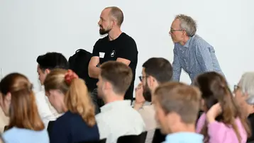 Zwei Männer stehen in einer Gruppe von Menschen. Der Mann links trägt ein schwarzes T-Shirt und schaut aufmerksam nach vorne, während der Mann rechts mit grauem Haar ebenfalls interessiert wirkt. Im Hintergrund sind mehrere Personen zu sehen, die dem Geschehen folgen.