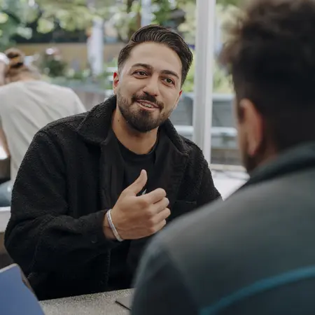 Zwei Männer sitzen an einem Tisch im Freien. Ein Mann mit dunklem, lockigem Haar spricht und zeigt mit der Hand ein Daumen-hoch-Zeichen. Der andere Mann, der ihm zuhört, hat die Rückenansicht zu sehen. Im Hintergrund sind unscharfe Pflanzen sichtbar.