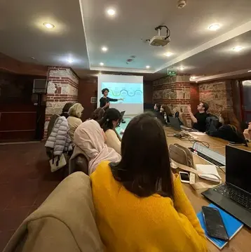 A speaker stands in front of a group of people in a meeting room at the UNESCO Chair workshop at RheinMain University of Applied Sciences in Bursa. In the background, a presentation showing a diagram can be seen on the wall. Those present are attentive, some taking notes. The room has a cozy atmosphere with bright lighting.