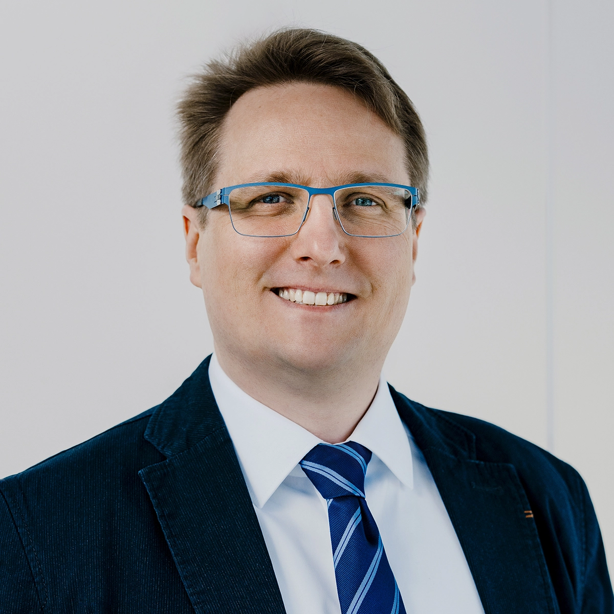 Ein lächelnder Mann mit Brille trägt einen Blazer und ein Hemd mit Krawatte. Der Hintergrund ist neutral und hell. Der Mann wirkt freundlich und professionell.
