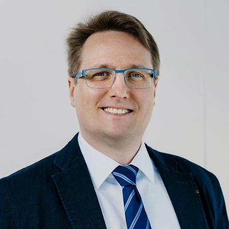 Ein lächelnder Mann mit Brille trägt einen Blazer und ein Hemd mit Krawatte. Der Hintergrund ist neutral und hell. Der Mann wirkt freundlich und professionell.
