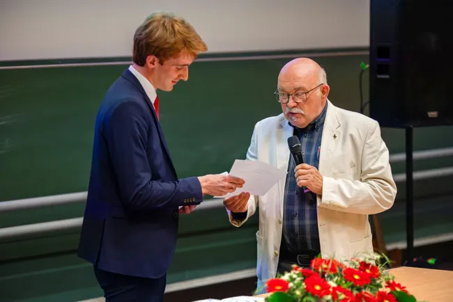 Ein junger Mann in Anzug überreicht ein Dokument an einen älteren Mann, der einen Mikrofon in der Hand hält. Beide stehen an einem Tisch, vor dem eine Blumenarrangement zu sehen ist. Der Hintergrund zeigt eine grüne Tafel und eine Lautsprecherbox.