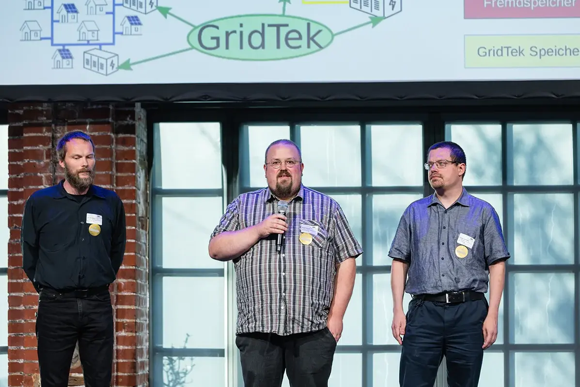 Drei Männer stehen auf einer Bühne und präsentieren ihr Projekt "GridTek"