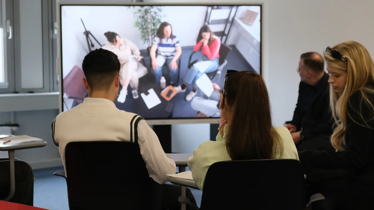 Eine Gruppe von Personen sitzt an einem Tisch und schaut auf einen großen Bildschirm, auf dem eine weitere Gruppe von Menschen zu sehen ist, die in einem Gespräch sind. Die Atmosphäre wirkt konzentriert und kommunikativ.