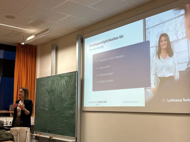 Die Präsentation zeigt eine Frau, die vor einem Bildschirm steht, auf dem Informationen über Einstiegsmöglichkeiten für Studierende bei Lufthansa Technik dargestellt sind. Die Punkte umfassen Praktika, Werkstudien, Abschlussarbeiten und duales Studium.