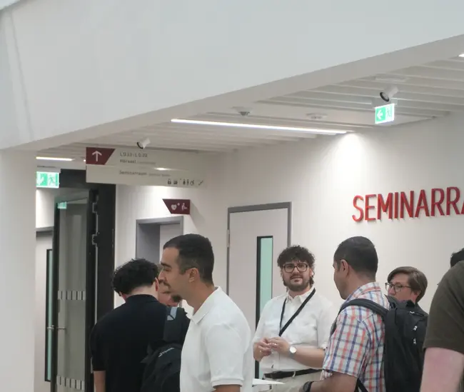 Gruppe von Personen in einem modernen Raum, die sich unterhalten. Einige tragen Rucksäcke, während andere entspannt stehen. Im Hintergrund ist ein Schild mit der Aufschrift "SEMINARRAUM" sichtbar. Die Atmosphäre wirkt informell und einladend.