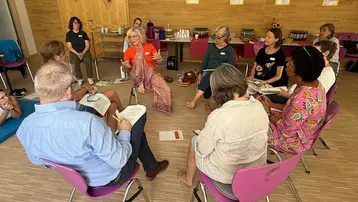 Eine Gruppe von Frauen und Männern sitzt in einem Kreis auf Stühlen und diskutiert lebhaft. In der Mitte steht eine Frau mit einem bunten Tuch und spricht. Im Hintergrund sind Tische mit Getränken und Materialien für die Veranstaltung zu sehen.