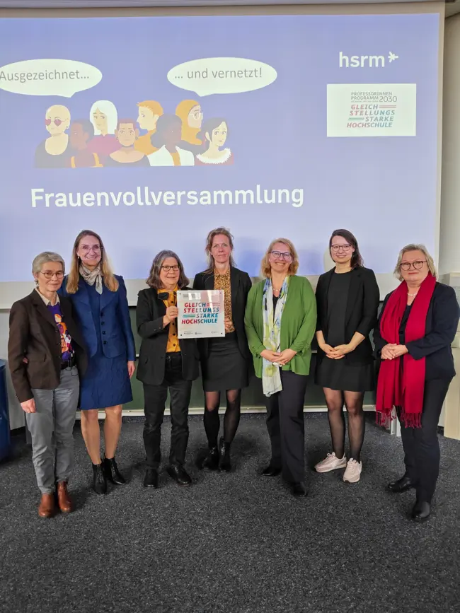 Eine Gruppe von sieben Frauen steht gemeinsam auf der Bühne, lächelt und hält ein Plakat. Im Hintergrund ist ein Banner mit der Aufschrift „Frauenvollversammlung“ und „Ausgezeichnet… und vernetzt!“ zu sehen. Die Frauen repräsentieren verschiedene Altersgruppen und Hintergründe.
