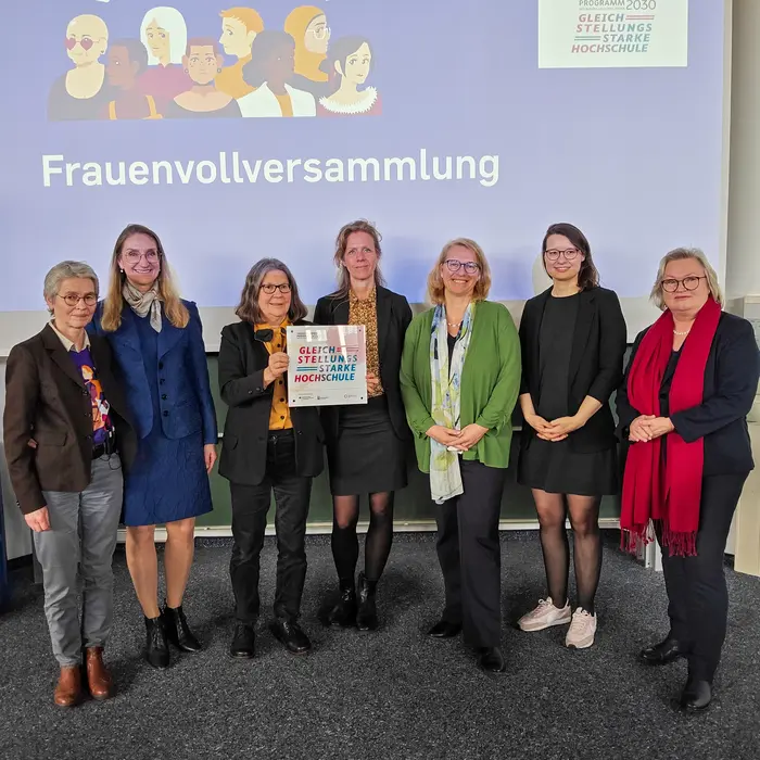 Eine Gruppe von sieben Frauen steht gemeinsam auf der Bühne, lächelt und hält ein Plakat. Im Hintergrund ist ein Banner mit der Aufschrift „Frauenvollversammlung“ und „Ausgezeichnet… und vernetzt!“ zu sehen. Die Frauen repräsentieren verschiedene Altersgruppen und Hintergründe.