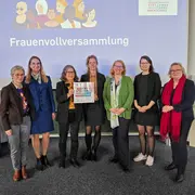 Eine Gruppe von sieben Frauen steht gemeinsam auf der Bühne, lächelt und hält ein Plakat. Im Hintergrund ist ein Banner mit der Aufschrift „Frauenvollversammlung“ und „Ausgezeichnet… und vernetzt!“ zu sehen. Die Frauen repräsentieren verschiedene Altersgruppen und Hintergründe.