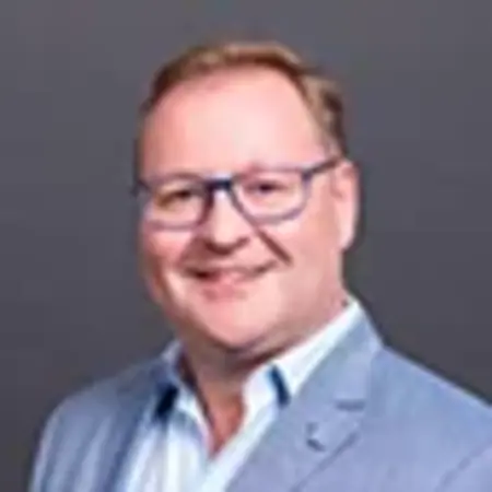 Ein lächelnder Mann mit Brille und blauer Jacke steht vor einem grauen Hintergrund. Er hat kurz geschnittenes, blondes Haar. Die positive Körpersprache vermittelt Freundlichkeit und Offenheit.