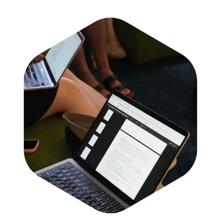 Eine Person sitzt mit einem Laptop auf den Knien, auf dem ein Dokument geöffnet ist. Der Bildschirm zeigt einen Text mit mehreren Punkten. Im Hintergrund sind Teile eines weiteren Laptops und Beine sichtbar.