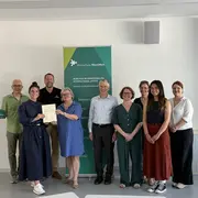Eine Gruppe von zehn Personen steht zusammen in einem Raum, einige halten Urkunden oder Dokumente. Sie lächeln und posieren vor einem Banner der Hochschule RheinMain, das internationale Zusammenarbeit thematisiert. Der Fokus liegt auf der feierlichen Atmosphäre und dem Austausch von Erfolgen.