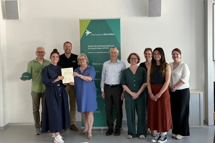 Eine Gruppe von zehn Personen steht zusammen in einem Raum, einige halten Urkunden oder Dokumente. Sie lächeln und posieren vor einem Banner der Hochschule RheinMain, das internationale Zusammenarbeit thematisiert. Der Fokus liegt auf der feierlichen Atmosphäre und dem Austausch von Erfolgen.