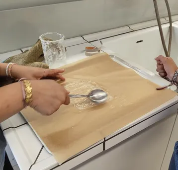 Eine Hand hält einen Löffel über eine braune Oberfläche, die mit einer glänzenden, transparenten Substanz bedeckt ist. Eine zweite Hand ist ebenfalls sichtbar, während im Hintergrund ein Glas und andere Küchenutensilien zu erkennen sind.