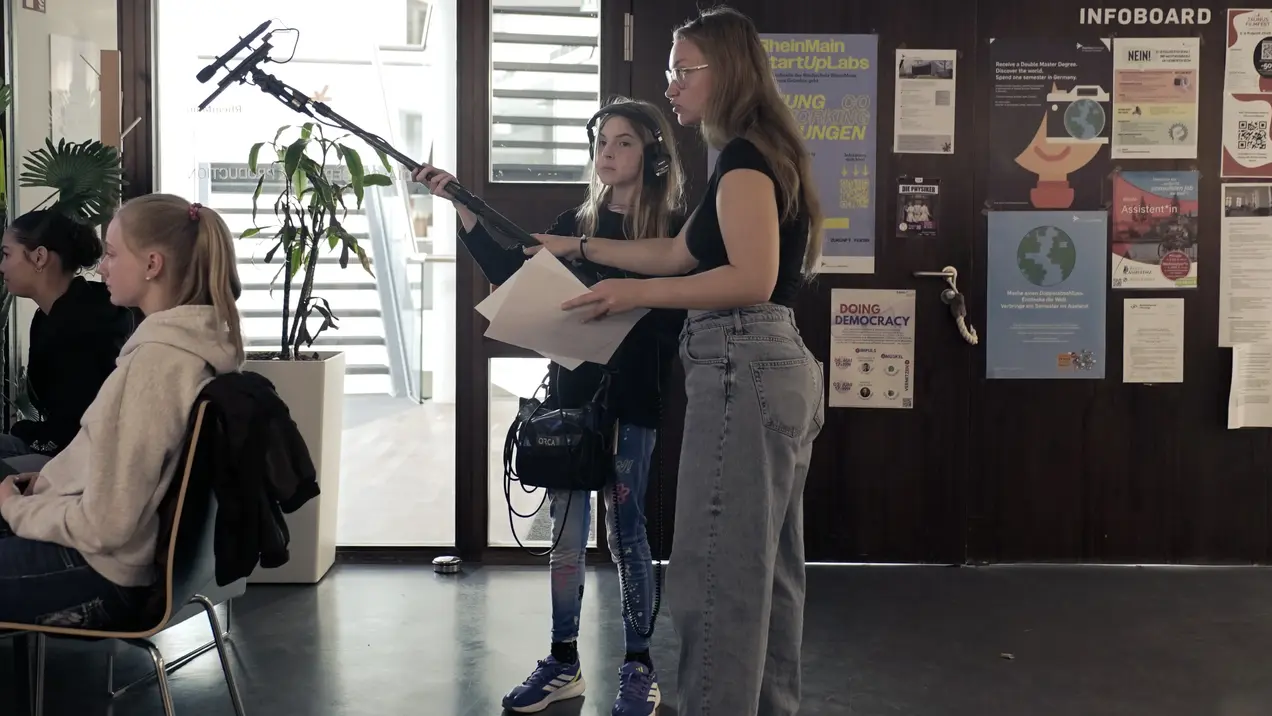 Zwei junge Frauen stehen in einem Raum. Eine hat ein Mikrofon und Notizen in der Hand, während die andere Kameraequipment trägt. Im Hintergrund sitzen Personen an Tischen. An der Wand hängen Flyer und Informationen. Die Szene vermittelt eine kreative oder filmische Arbeitsatmosphäre.