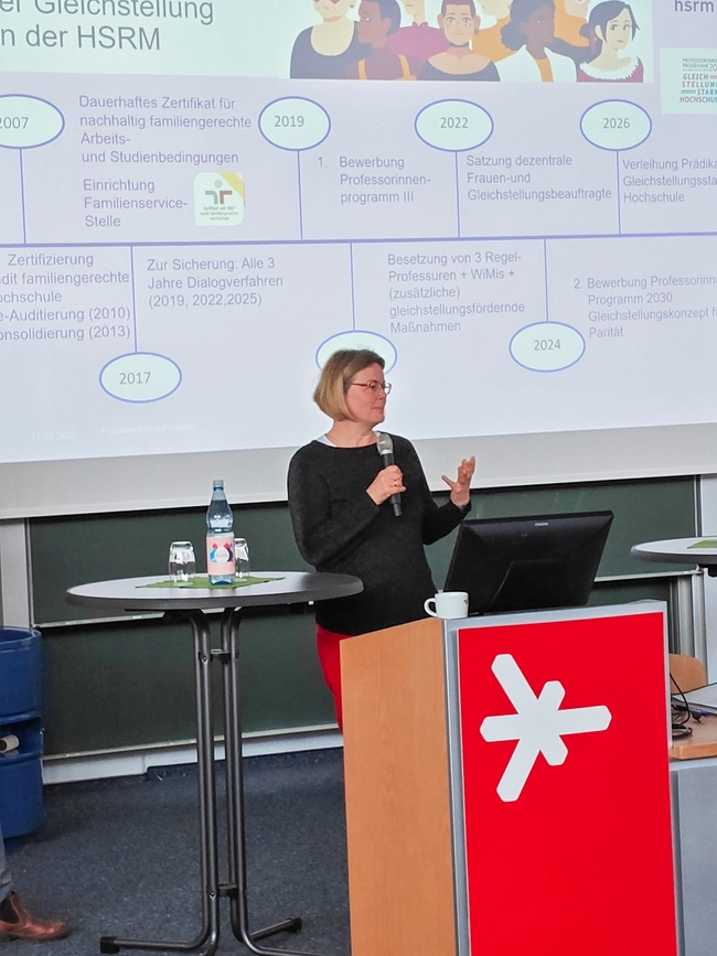 Eine Frau steht an einem Rednerpult und präsentiert Informationen zur Gleichstellung an der Hochschule. Im Hintergrund sind eine Pinnwand mit Zeitstrahl und verschiedene Meilensteine zur Gleichstellung und Zertifizierungsprozessen zu sehen. Auf dem Tisch stehen Wasserflaschen.