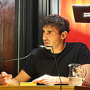 Poetikdozent Necati Öziri sitzt an einem Tisch und liest aus seinem Buch, das vor ihm liegt