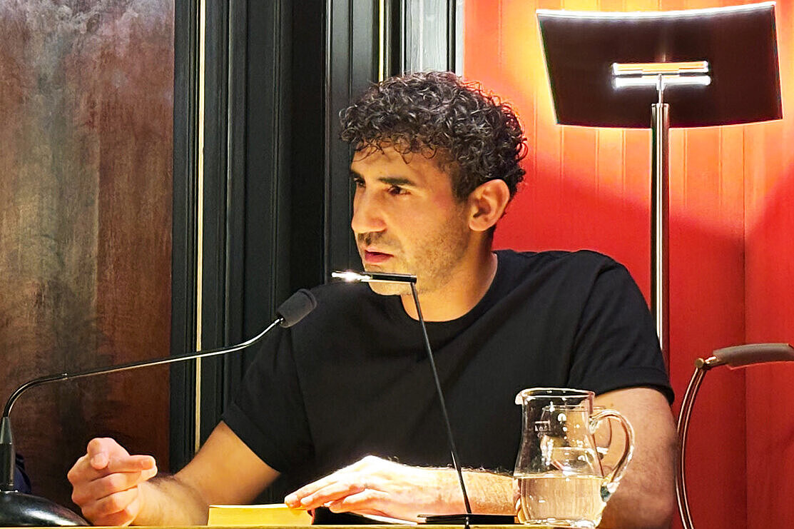 Poetikdozent Necati Öziri sitzt an einem Tisch und liest aus seinem Buch, das vor ihm liegt