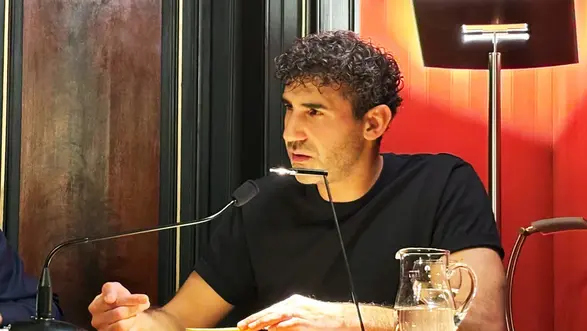 Poetikdozent Necati Öziri sitzt an einem Tisch und liest aus seinem Buch, das vor ihm liegt