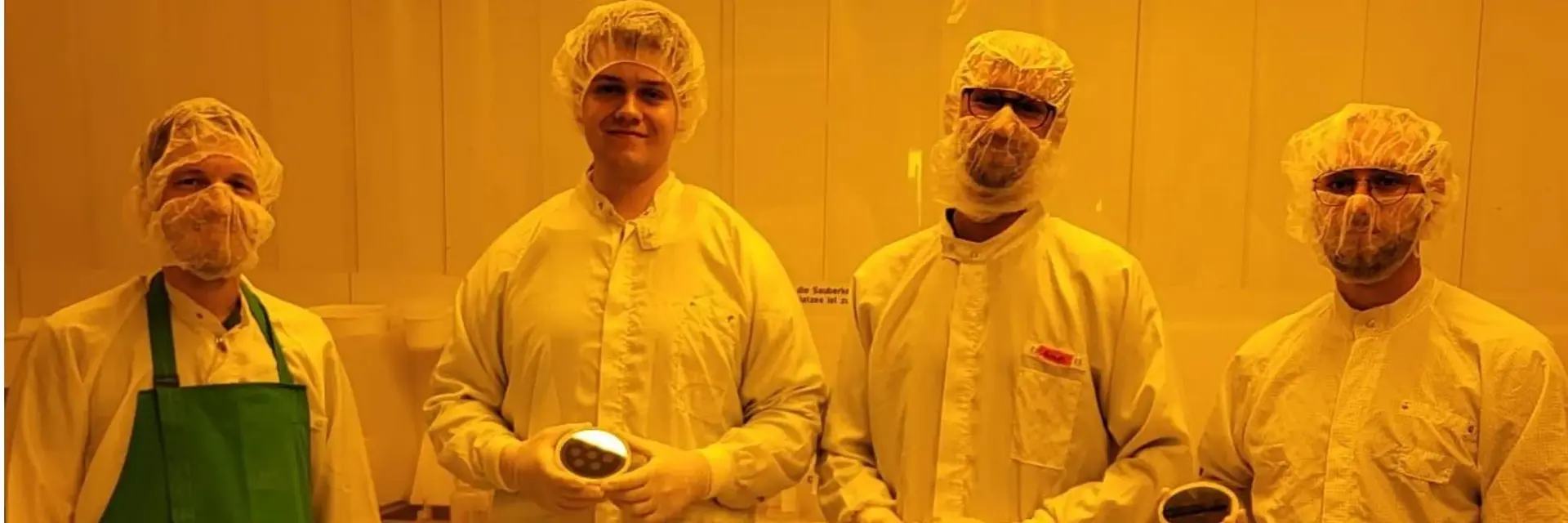 Vier Personen in Schutzkleidung stehen zusammen in einem Labor mit gelblich-orangefarbener Beleuchtung. Sie tragen Haarnetze, Mundschutz und Schutzbrillen. Jede Person hält ein kleines Objekt in der Hand. Die Atmosphäre ist sauber und professionell, typisch für einen technischen oder wissenschaftlichen Bereich.