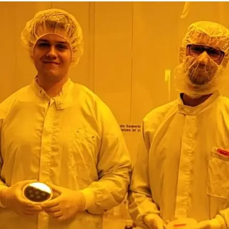 Vier Personen in Schutzkleidung stehen zusammen in einem Labor mit gelblich-orangefarbener Beleuchtung. Sie tragen Haarnetze, Mundschutz und Schutzbrillen. Jede Person hält ein kleines Objekt in der Hand. Die Atmosphäre ist sauber und professionell, typisch für einen technischen oder wissenschaftlichen Bereich.