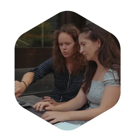 Zwei Frauen sitzen am Tisch und schauen konzentriert auf einen Laptop. Eine der Frauen zeigt auf den Bildschirm, während die andere aufmerksam zuschaut. Sie scheinen an einem Projekt oder einer Aufgabe zu arbeiten.