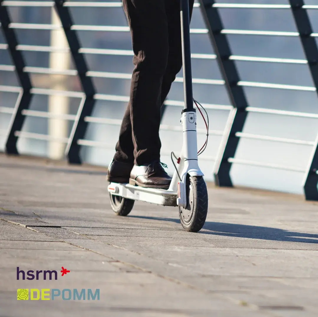 Eine Person fährt auf einem E-Scooter entlang einer gepflasterten Fläche. Im Hintergrund ist ein Geländer sichtbar, das an einem Gewässer entlangführt. Der Fokus liegt auf dem E-Scooter und der Person, die in Bewegung sind.