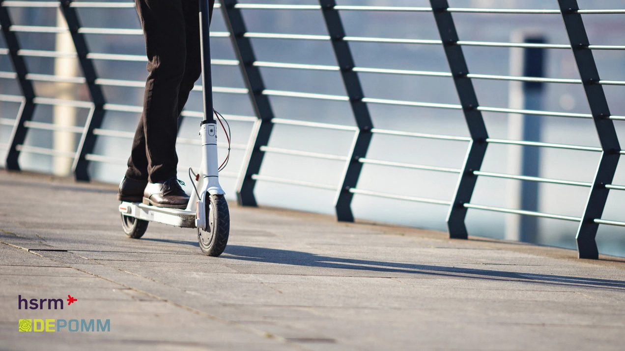 Eine Person fährt auf einem E-Scooter entlang einer gepflasterten Fläche. Im Hintergrund ist ein Geländer sichtbar, das an einem Gewässer entlangführt. Der Fokus liegt auf dem E-Scooter und der Person, die in Bewegung sind.