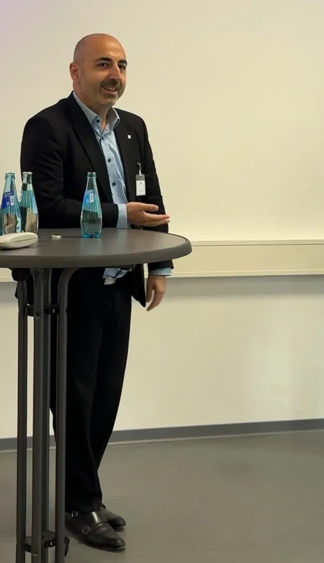 Ein Mann in Anzug steht an einem Stehtisch und hält ein Smartphone in der Hand. Auf dem Tisch sind mehrere Flaschen Wasser sichtbar. Die Umgebung sieht modern und leicht nüchtern aus, mit weißen Wänden und grauem Boden. Der Mann spricht mit einem freundlichen Ausdruck.