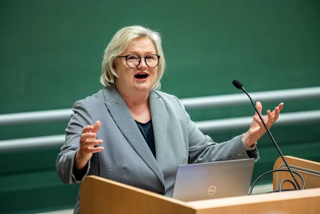 Eine Frau mit blonden, schulterlangen Haaren steht an einem Rednerpult und spricht leidenschaftlich. Sie trägt einen karierten Blazer und hat eine begeisterte Gestik. Im Hintergrund ist eine grüne Wand zu sehen.