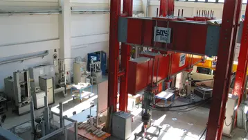 Eine große industrielle Testanlage mit roten und grauen Strukturen. Im Inneren gibt es verschiedene Maschinen und Labortische. Das Licht fällt durch große Fenster auf den Boden und die Geräte, die für Tests und Experimente verwendet werden.