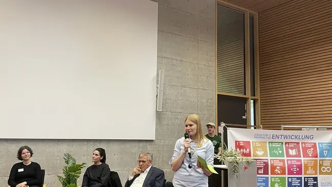 Eine Frau mit blonden Haaren steht am Rednerpult und hält einen Vortrag. Im Hintergrund sitzen mehrere Personen auf Stühlen. An der Wand ist eine große Leinwand mit Symbolen für nachhaltige Entwicklung zu sehen. Der Raum ist hell und modern gestaltet.