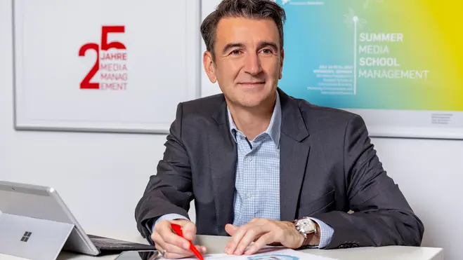 Ein Mann in Anzug sitzt an einem Tisch mit einem Tablet und einer Stift in der Hand, während er auf Dokumente vor ihm schaut. Im Hintergrund sind Plakate mit den Aufschriften "25 Jahre Media Management" und "Summer Media School Management" zu sehen.