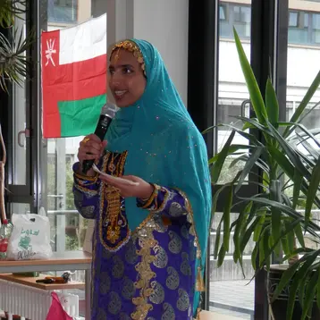 Eine Frau in traditioneller ausländischer Kleidung spricht lächelnd ins Mikrofon. Sie trägt einen bunten, schimmernden Anzug mit goldenen Verzierungen und einen passenden Hijab. Im Hintergrund sind Pflanzen und eine Fahne sichtbar, die eine Veranstaltung umrahmen.