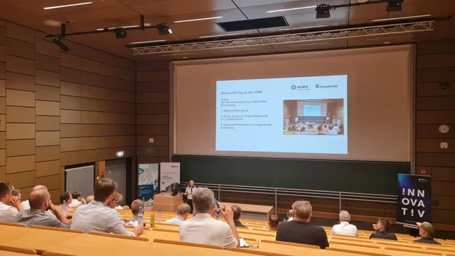 Ein Vortrag in einem Seminarraum mit Reihen von Sitzen. Auf einer Leinwand wird eine Präsentation gezeigt, die Informationen über ein Projekt oder eine Veranstaltung enthält. Im Vordergrund sitzt ein Redner, während das Publikum aufmerksam zuhört.
