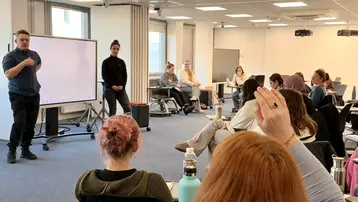 In einem Klassenzimmer stehen zwei Personen vorne; eine spricht, während die andere aufmerksam zuhört. Im Publikum hebt eine Person die Hand, um eine Frage zu stellen. Die Atmosphäre wirkt interaktiv und lehrreich.