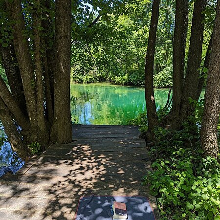 Entspannt lesen am Fluss in Bellavici, Kroatien Es ist ein Holzsteg zu sehen, auf dem ein Handtuch mit Buch und HSRM-Tasche liegt. Ein türkiser Fluss im Hintergrund schimmert in der Sonne