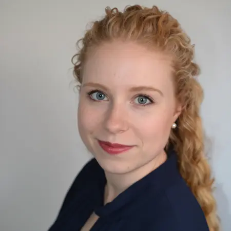 Ein Porträt einer jungen Frau mit lockigem, blondem Haar und blauen Augen. Sie trägt ein dunkles Oberteil und hat einen sanften, freundlichen Ausdruck. Der Hintergrund ist neutral, um den Fokus auf ihr Gesicht zu lenken.