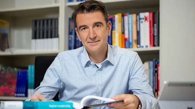 Ein Mann mit dunklen Haaren sitzt an einem Tisch, hält ein Dokument in der Hand und schaut freundlich in die Kamera. Im Hintergrund sind Bücherregale mit verschiedenen Büchern sichtbar. Auf dem Tisch liegen einige Bücher, darunter ein Exemplar mit dem Titel "digital copyright".