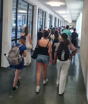 Eine Gruppe von Jugendlichen und jungen Erwachsenen geht in einem Flur. Sie tragen verschiedene Freizeitkleidung und Rucksäcke. Einige Personen sind sichtbar mit Smartphones beschäftigt. Der Flur hat große Fenster, die Tageslicht hereinlassen.