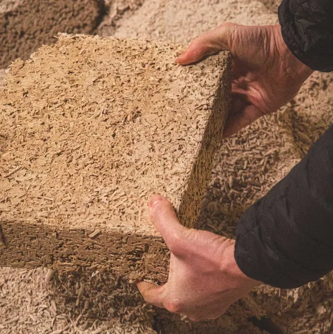 Eine Person hält einen quadratischen Block aus Holzmaterial in beiden Händen. Der Block wird aus einem Boden von Holzspänen und -abfällen herausgehoben. Die Szene vermittelt einen Eindruck von handwerklicher Arbeit mit natürlichen Materialien.