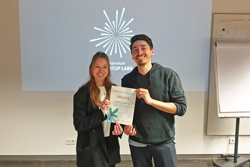 Die Kommunikationsdesign-StudierendenCharlotte Fischer und Marcel Beck stehen mit der Urkunde für den 2. Platz vor ihrem Logoentwurf