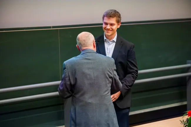 Zwei Männer stehen in einem Raum mit einer grünen Wand. Einer von ihnen lächelt und scheint einen Preis oder eine Auszeichnung zu erhalten, während der andere ihm gegenübersteht und ihn anspricht. Es herrscht eine freundliche Atmosphäre.