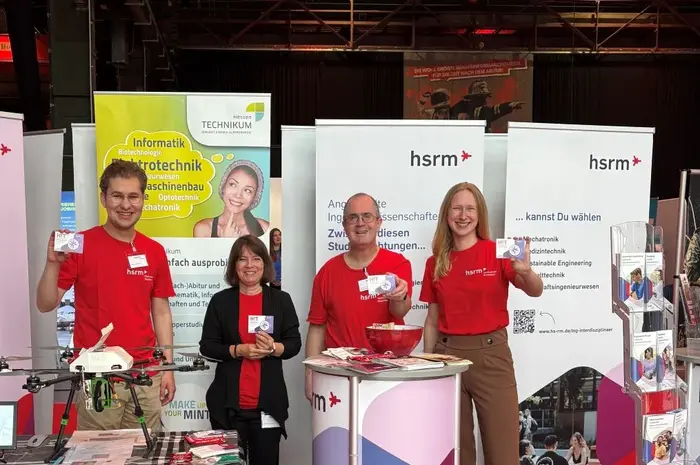 Auf dem Bild sind vier Personen in roten T-Shirts am Stand der Hochschule für Technik und Wirtschaft. Sie lächeln und halten Informationsmaterial in der Hand. Im Hintergrund sind Banner mit Informationen zur Hochschule zu sehen. Der Stand ist beleuchtet und freundlich gestaltet.