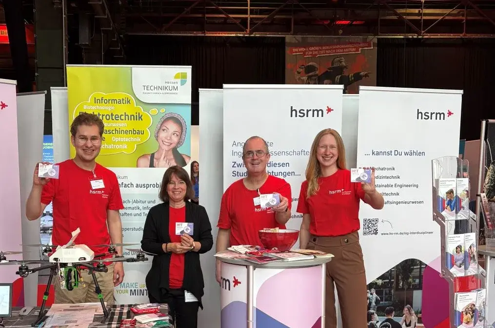 Auf dem Bild sind vier Personen in roten T-Shirts am Stand der Hochschule für Technik und Wirtschaft. Sie lächeln und halten Informationsmaterial in der Hand. Im Hintergrund sind Banner mit Informationen zur Hochschule zu sehen. Der Stand ist beleuchtet und freundlich gestaltet.